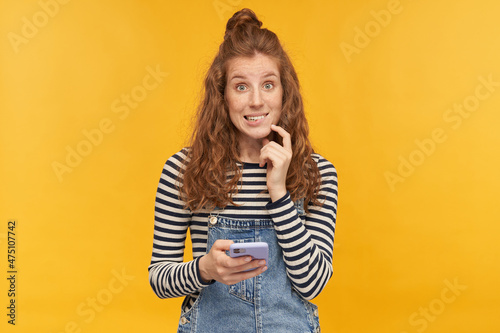 indoor studio shot of young ginger female with long red curly hair biting her lip, holding phone with ashamed guilty facial expression, isolated over yellow background