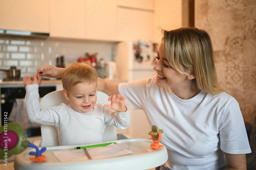 Little cute baby toddler boy blonde sitting on baby chair learning to draw. Beautiful young mom and son playing spend time together indoors at home with toys. Healthy happy family childhood concept Little cute baby toddler boy blonde sitting on baby chair learning to draw. Beautiful young mom and son playing spend time together indoors at home with toys. Healthy happy family childhood concept
