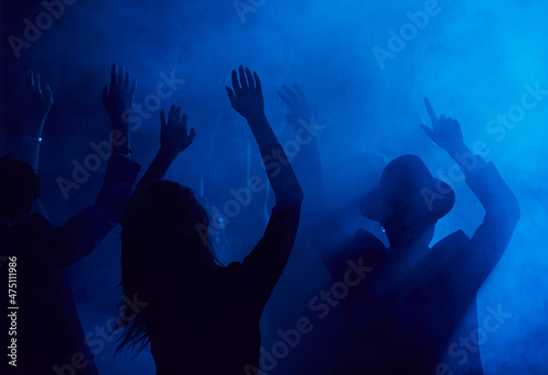 Silhouettes of young people jumping and raising hands while enjoying party in smoky nightclub lit by blue light, copy space