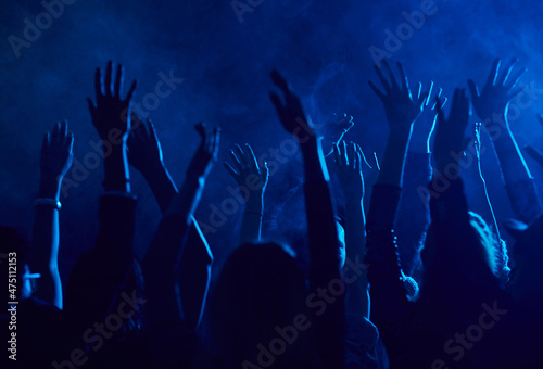 Silhouette of large group of people raising hands while enjoying music concert in smoky nightclub lit by blue light, copy space