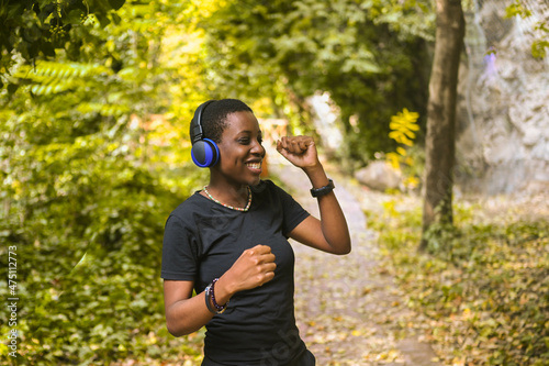 Attractive happy smiling young natural beauty short haired African Black woman with blue headphones in black t-shirt listening music and dancing in nature summer park