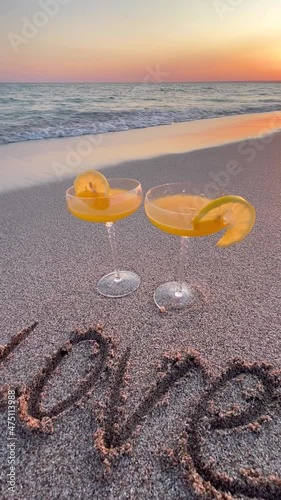 Two glasses of orange cocktail and the word Love on the sea sand at sunset