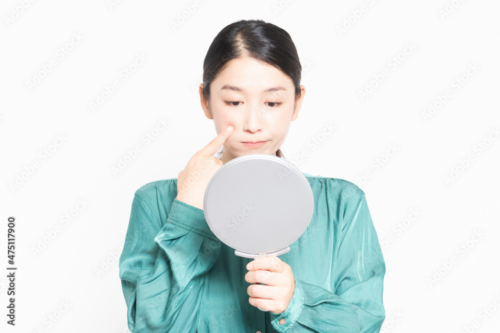 白い背景の前に立って、鏡で肌のチェックをする主婦