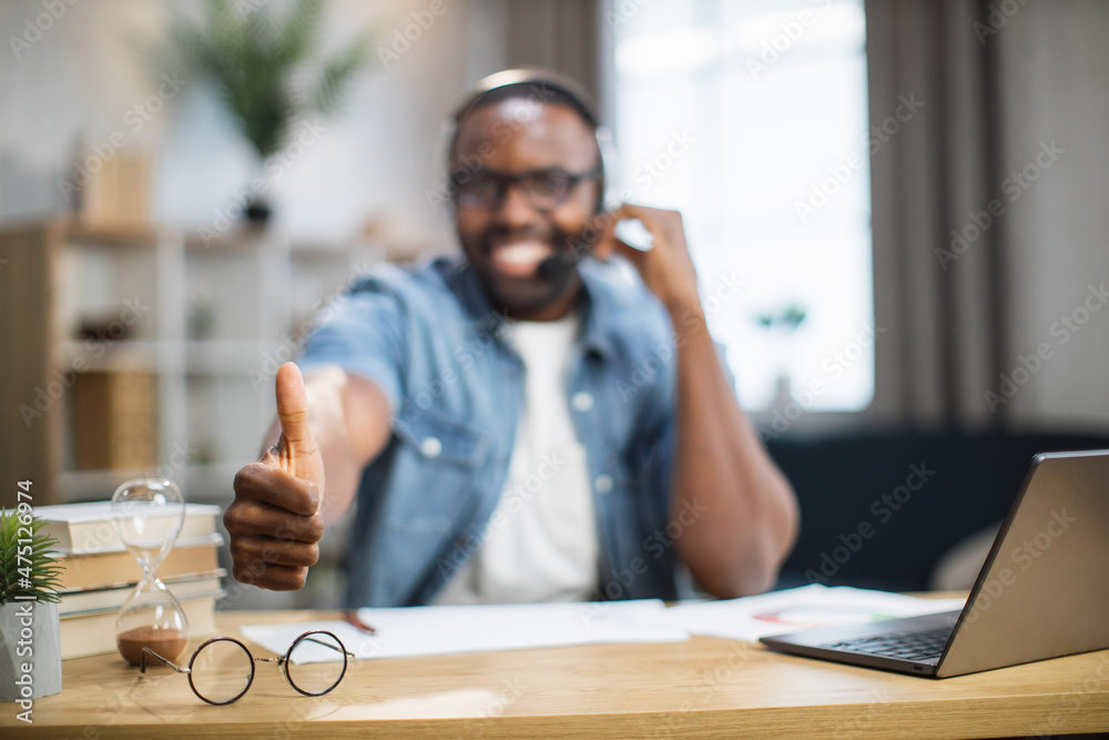 Blur view of smiling african american man in headset showing thumb up on camera while sitting at desk with modern laptop. Happy male freelancer at home. Focus on eyeglasses on the table Blur view of smiling african american man in headset showing thumb up on camera while sitting at desk with modern laptop. Happy male freelancer at home. Focus on eyeglasses on the table