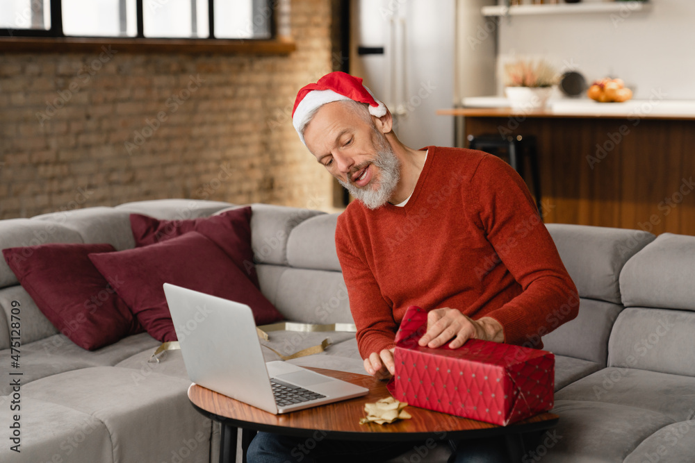 Curious surprised caucasian mature middle-aged man in Santa Claus red hat celebrating Christmas New Year online while having video call unpacking unwrapping present gift box at home Curious surprised caucasian mature middle-aged man in Santa Claus red hat celebrating Christmas New Year online while having video call unpacking unwrapping present gift box at home