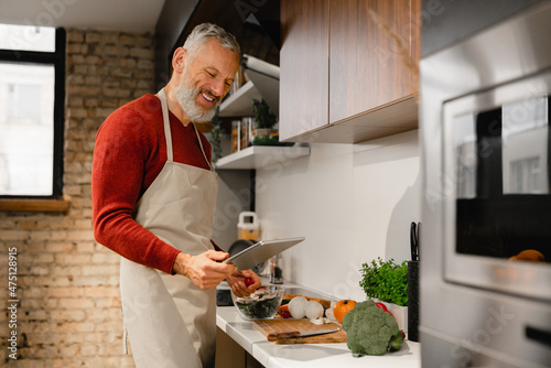 Mature middle-aged man chef cooking, looking for recipe on digital tablet online, preparing vegetable vegan vegetarian salad food meal at home kitchen