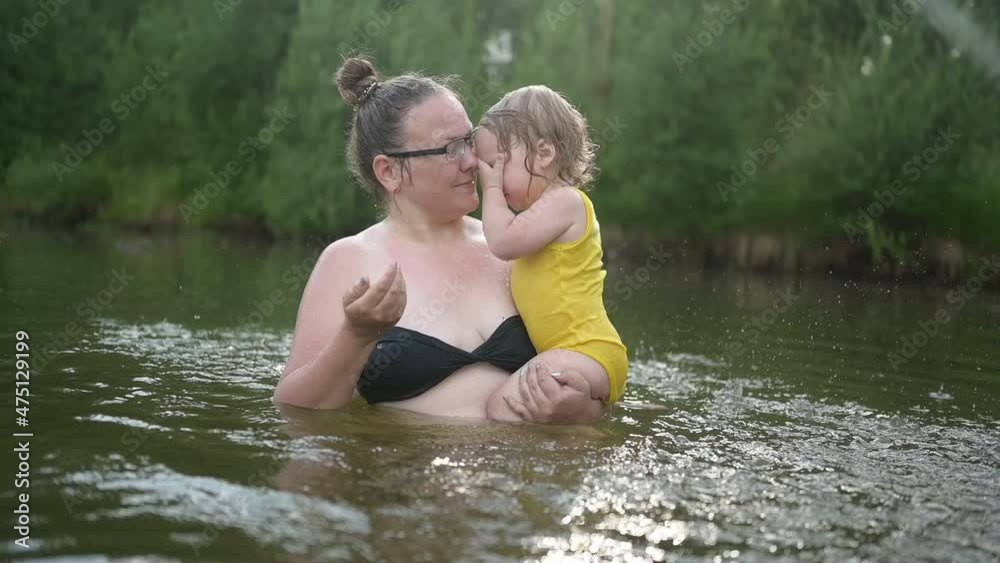 Little funny cute blonde girl child toddler in yellow bodysuit laughing learns swim outside at summer lake. Plus size body positive smiling woman mother baby swimming in natural pool. Water splashes Little funny cute blonde girl child toddler in yellow bodysuit laughing learns swim outside at summer lake. Plus size body positive smiling woman mother baby swimming in natural pool. Water splashes
