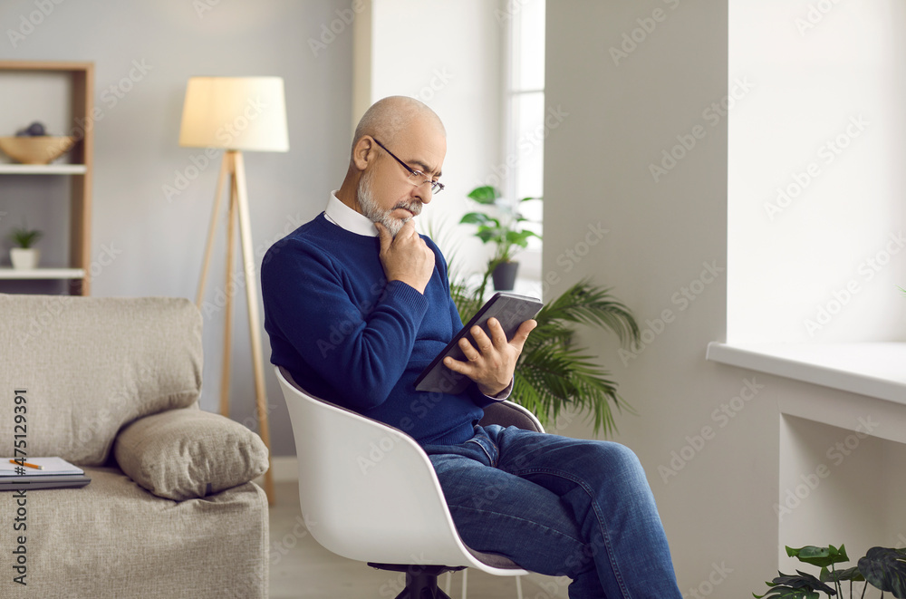 Senior man using a modern tablet computer. Serious mature man sitting in a comfortable chair at home, holding his tablet, reading an ebook, scratching his chin and thinking