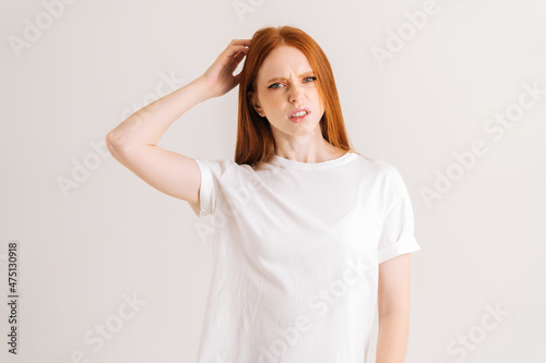 Studio portrait of bewildered pretty young woman pondering over something touching head standing on white isolated background. Front view of redhead stressed lady scratching head as seeks. © dikushin