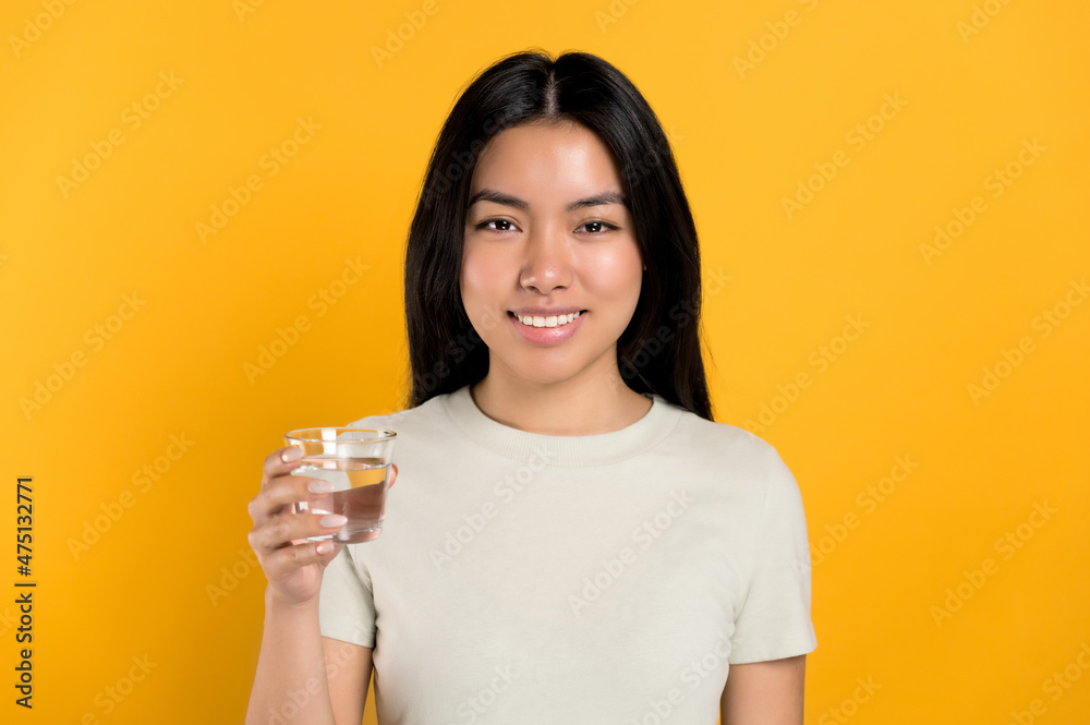 Positive, lovely chinese young woman in a t-shirt, holds a glass of clean water, cares of her health, drinks the daily norm of water, stands on isolated orange background. Healthy lifestyle concept