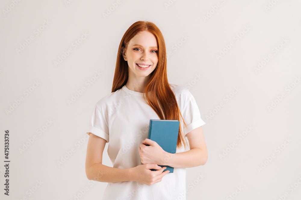 Studio portrait of cheerful young student woman holding in hands paper book and looking at camera, standing on white isolated background. Happy redhead lady posing with educational materials. Studio portrait of cheerful young student woman holding in hands paper book and looking at camera, standing on white isolated background. Happy redhead lady posing with educational materials.