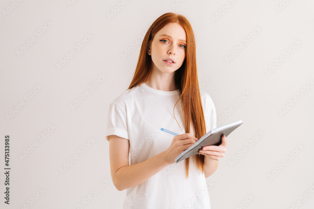 Studio portrait of pretty redhead young woman writing in notebook looking at camera, standing on white isolated background. Confident female student making notes in paper diary.