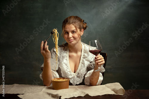Half-length portrait of young beautiful girl in gray dress of medieval style having dinner isolated on dark vintage background.