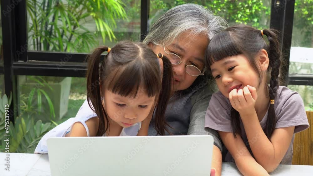 Asian Grandmother with her two grandchildren having fun and playing education games online with a computer notebook at home in the living room. Concept of online education and caring from parents. Asian Grandmother with her two grandchildren having fun and playing education games online with a computer notebook at home in the living room. Concept of online education and caring from parents.