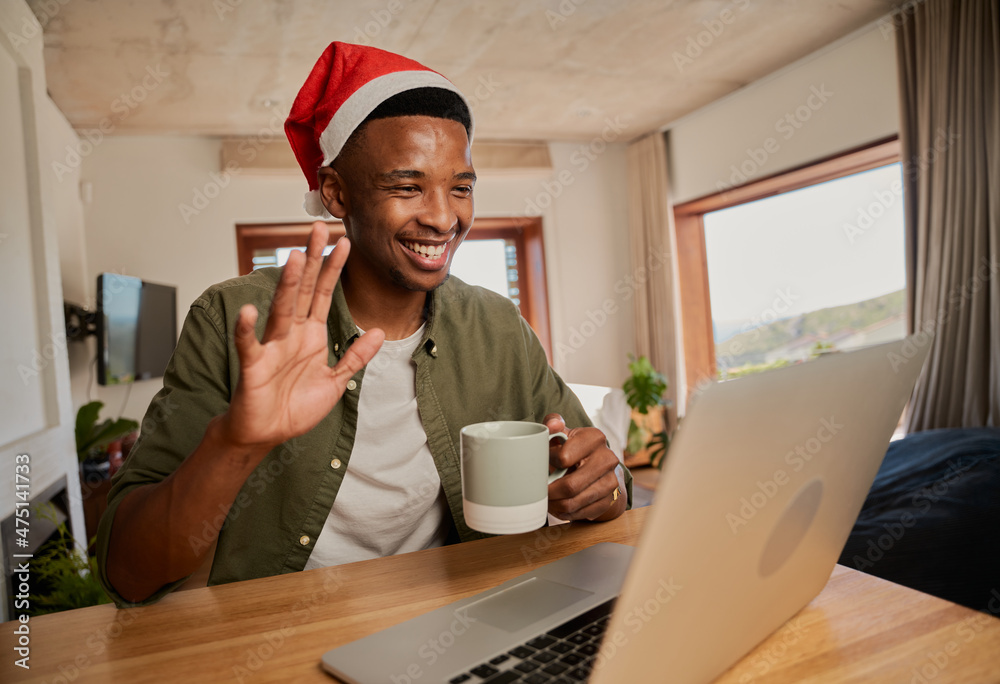 Young adult black male wearing Christmas hat, waving while on online meeting. Sitting at kitchen counter using laptop. Young adult black male wearing Christmas hat, waving while on online meeting. Sitting at kitchen counter using laptop.