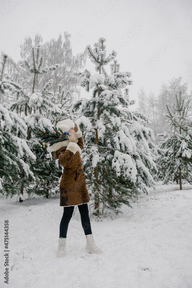A girl in a sheepskin coat, mittens, a white hat with earflaps and boots stands around snow-covered pine trees with a small artificial tree dressed up with New Year's toys and looks away