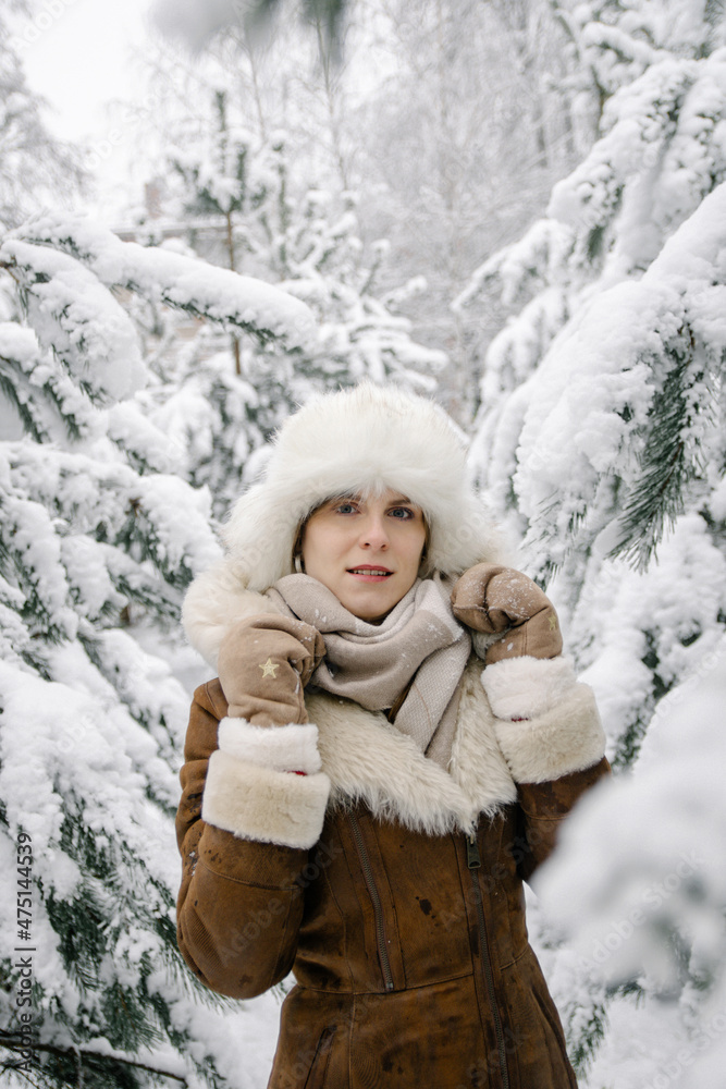 Vertical portrait of a smiling young european girl in a sheepskin coat, mittens, a white hat with earflaps and boots, who stands in snowy pine trees in winter Vertical portrait of a smiling young european girl in a sheepskin coat, mittens, a white hat with earflaps and boots, who stands in snowy pine trees in winter