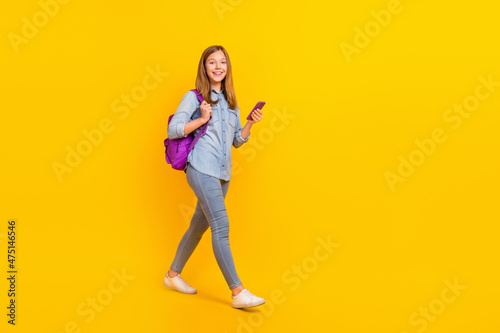 Full size photo of funny little girl go look telephone wear bag jeans shirt sneakers isolated on yellow background