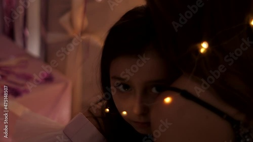 girl lies on her mother's shoulder tied with a garland and looks into the camera at night on Christmas day