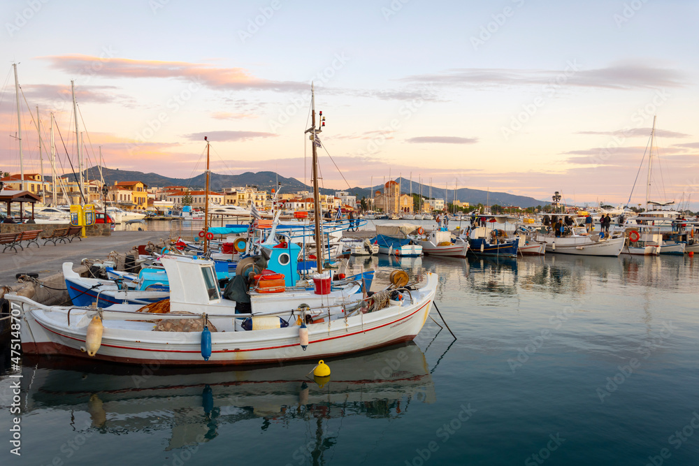 Colorful fishing boats line the harbor of the Greek island of Aegina, Greece at dusk, with the waterfront promenade and shops in view.