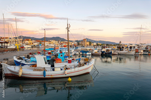 Colorful fishing boats line the harbor of the Greek island of Aegina, Greece at dusk, with the waterfront promenade and shops in view.