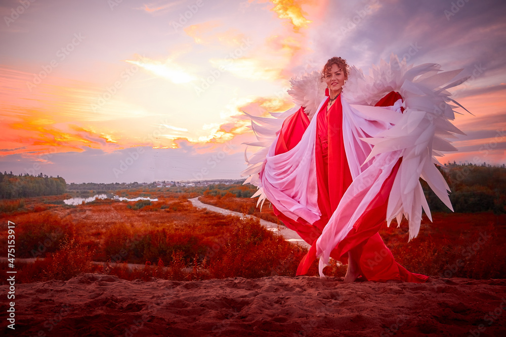Beautiful young woman or girl with curly hair and in red dress with a light flying fabric and white wings on the sand on sunny day with blue sky. Angel model or dancer posing in photo shoot on dunes