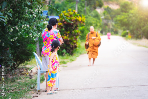 Mother and daughter stand and wait monks to walk alms on road in front house in order to make merit. In rural area with many trees. On morning time.