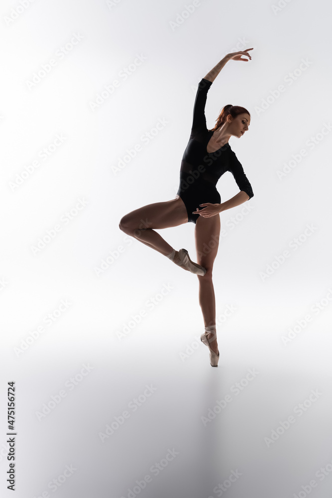 full length of young ballerina in pointe shoes and black bodysuit dancing on grey