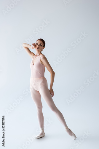 full length of elegant ballerina in bodysuit looking at camera on grey