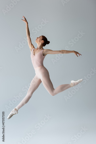 full length of graceful ballerina in bodysuit levitating and gesturing on grey