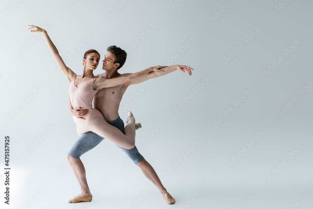 full length of shirtless man and young woman in bodysuit performing ballet dance on grey