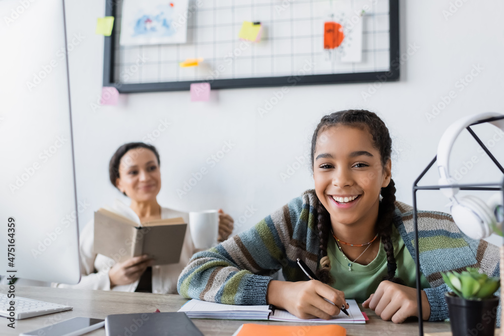 happy african american girl looking at camera while writing in notebook near mother with book on blurred background happy african american girl looking at camera while writing in notebook near mother with book on blurred background