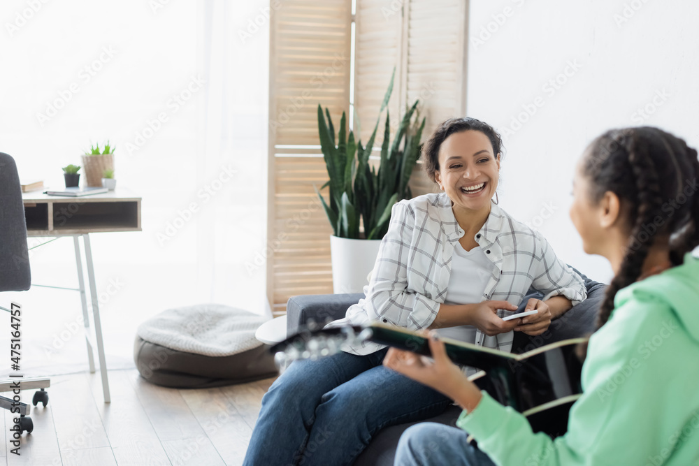 blurred african american girl playing guitar near laughing mom sitting on couch with cellphone blurred african american girl playing guitar near laughing mom sitting on couch with cellphone