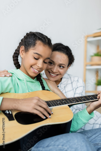 african american teenage girl playing acoustic guitar near happy mom at home