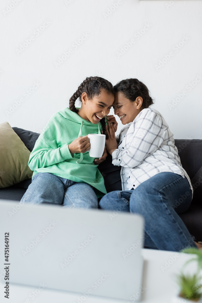 excited african american girl with cup of tea laughing together with mom while watching movie on blurred laptop