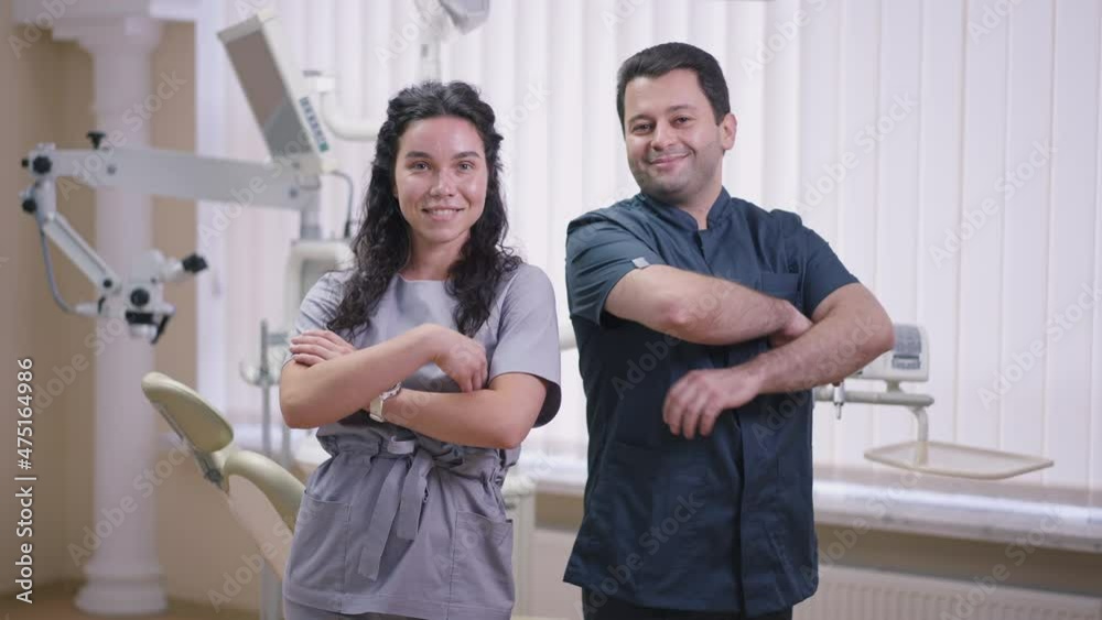 Male dentist and female assistant crossing hands smiling looking at camera with confident facial expression. Medium shot portrait Middle Eastern man and Caucasian woman posing in slow motion Male dentist and female assistant crossing hands smiling looking at camera with confident facial expression. Medium shot portrait Middle Eastern man and Caucasian woman posing in slow motion
