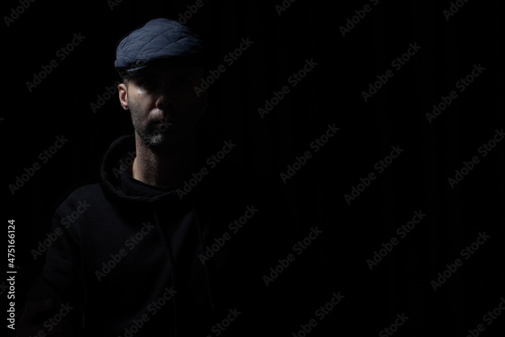 man in kepi and black hoodie is staying in darkness, light and shadows on his face man in kepi and black hoodie is staying in darkness, light and shadows on his face