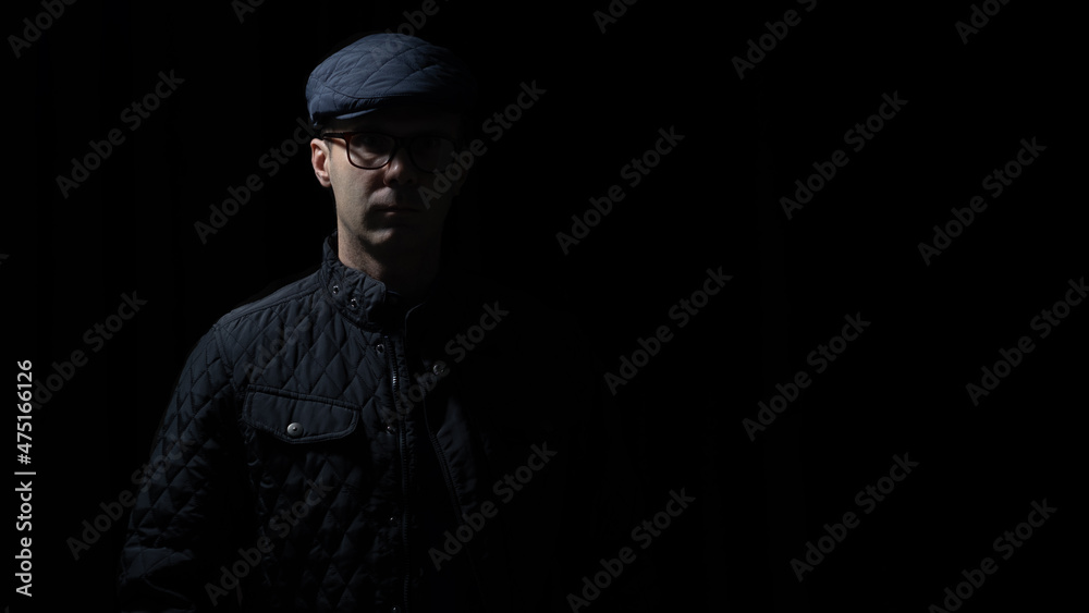 man in glasses, kepi and black jacket is staying in darkness, light and shadows on his face man in glasses, kepi and black jacket is staying in darkness, light and shadows on his face