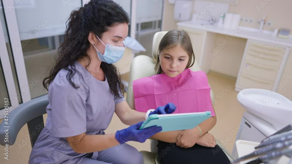 Cute curios teenage girl sitting in dentist chair listening to professional doctor showing tablet talking. Portrait of interested Caucasian teenager visiting orthodontist in hospital indoors. Slow Cute curios teenage girl sitting in dentist chair listening to professional doctor showing tablet talking. Portrait of interested Caucasian teenager visiting orthodontist in hospital indoors. Slow
