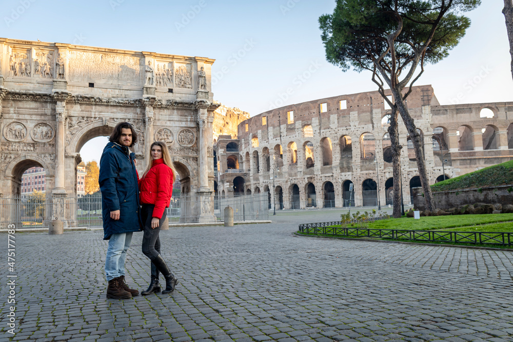 Young couple traveling to Rome. The young couple poses in front of the Arch of Titus. The blonde woman in her red jacket puts her hand on her boyfriend.