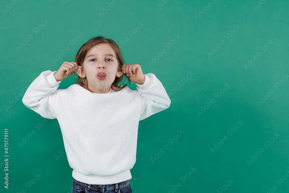 a boy with long hair makes faces, shows his tongue and holds his ears on a green isolated background. children's joy