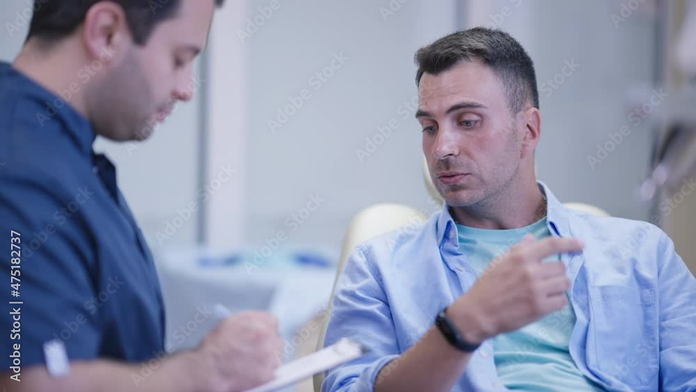 Portrait of handsome Caucasian man talking with Middle Eastern doctor sitting in dentist chair in hospital. Worried male patient complaining toothache indoors in slow motion. Orthodontist visit Portrait of handsome Caucasian man talking with Middle Eastern doctor sitting in dentist chair in hospital. Worried male patient complaining toothache indoors in slow motion. Orthodontist visit