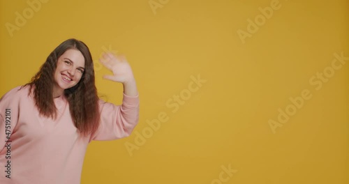 Smiling young girl blogger greeting subscribers wave hand say hello on yellow background with copy space for advertising