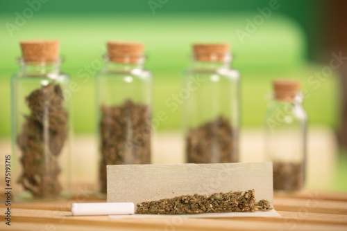 chopped marijuana, on cigarette paper with a filter and airtight glass jars with different types of cannabis on wood, process for the production of marijuana cigars. green background.