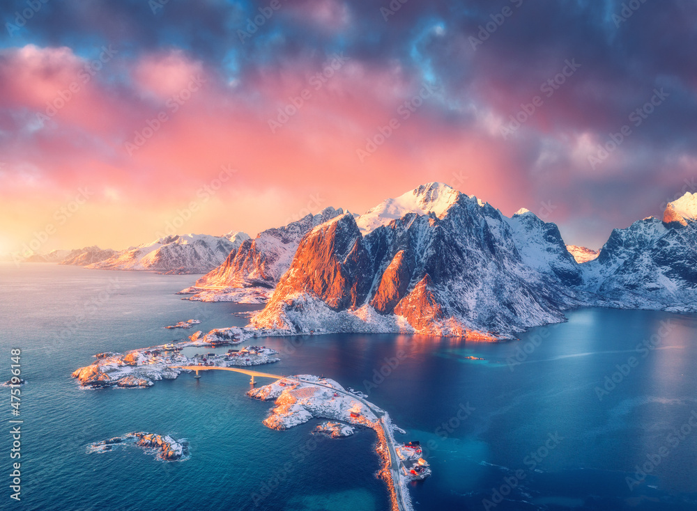 Beautiful landscape with blue sea, snowy mountains, rocks and islands, village, rorbu, road, bridge and pink sky at sunrise. Aerial view. Hamnoy in snow in winter in Lofoten islands, Norway. Top view - obrazy, fototapety, plakaty 