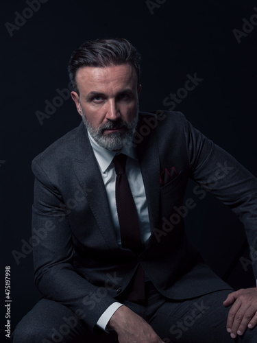 Portrait of adult businessman wearing trendy suit and sitting in modern studio on stylish chair against the black background