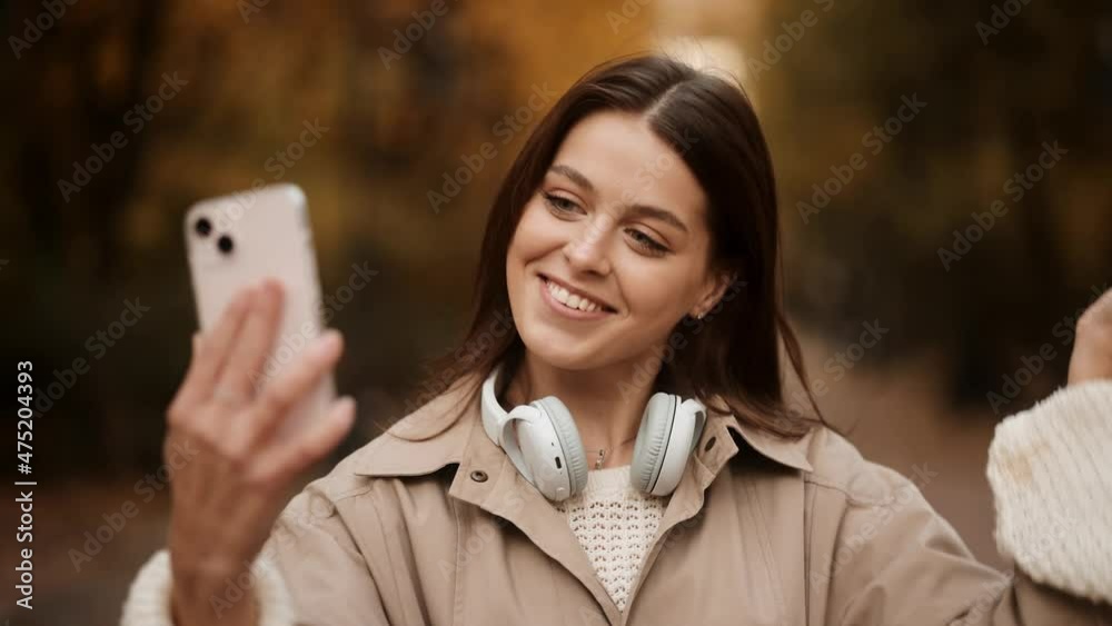 A young woman in a white sweater and trench coat answers a video call. The concept of online communication. Attractive brunette woman walking in the park at fall on a sunny day and smiling