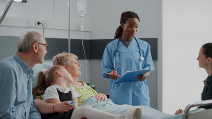 Close up of medical assistant explaining disease to patient and family in visit in hospital ward. Woman nurse talking about treatment and recovery to visitors and retired person with sickness.
