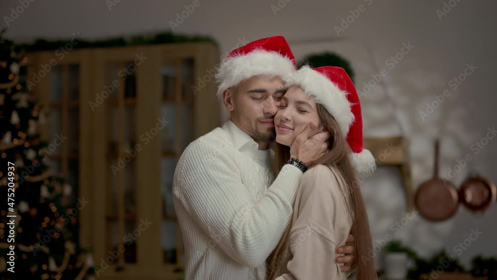 Portrait Of Young People In Santa Hats. Young Man Holds In His Arms Pretty Girl With Long Hair. They Kiss.