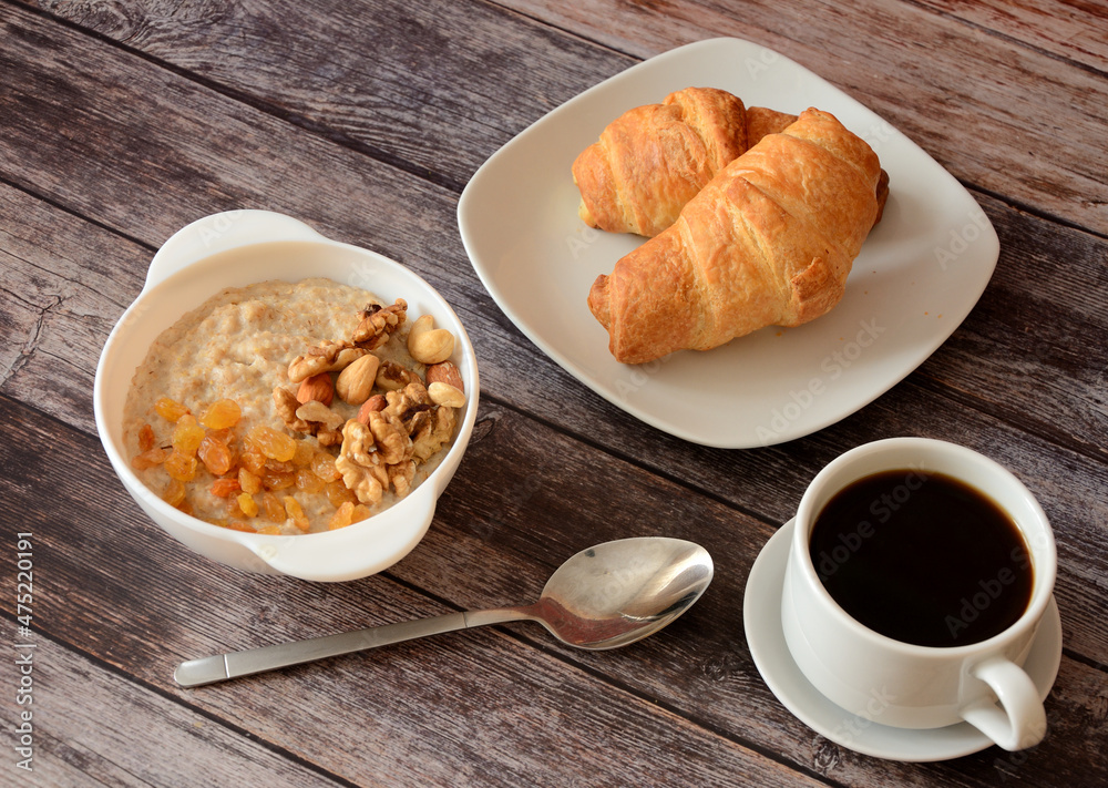 Delicious breakfast, a cup of oatmeal with raisins and nuts, two croissants and a cup of black coffee on a wooden table.
